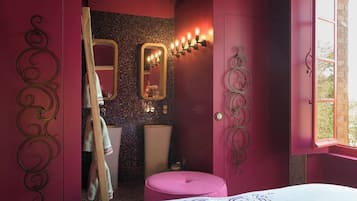 A room with a bed, a pink ottoman, a mirror with lights, and a window.