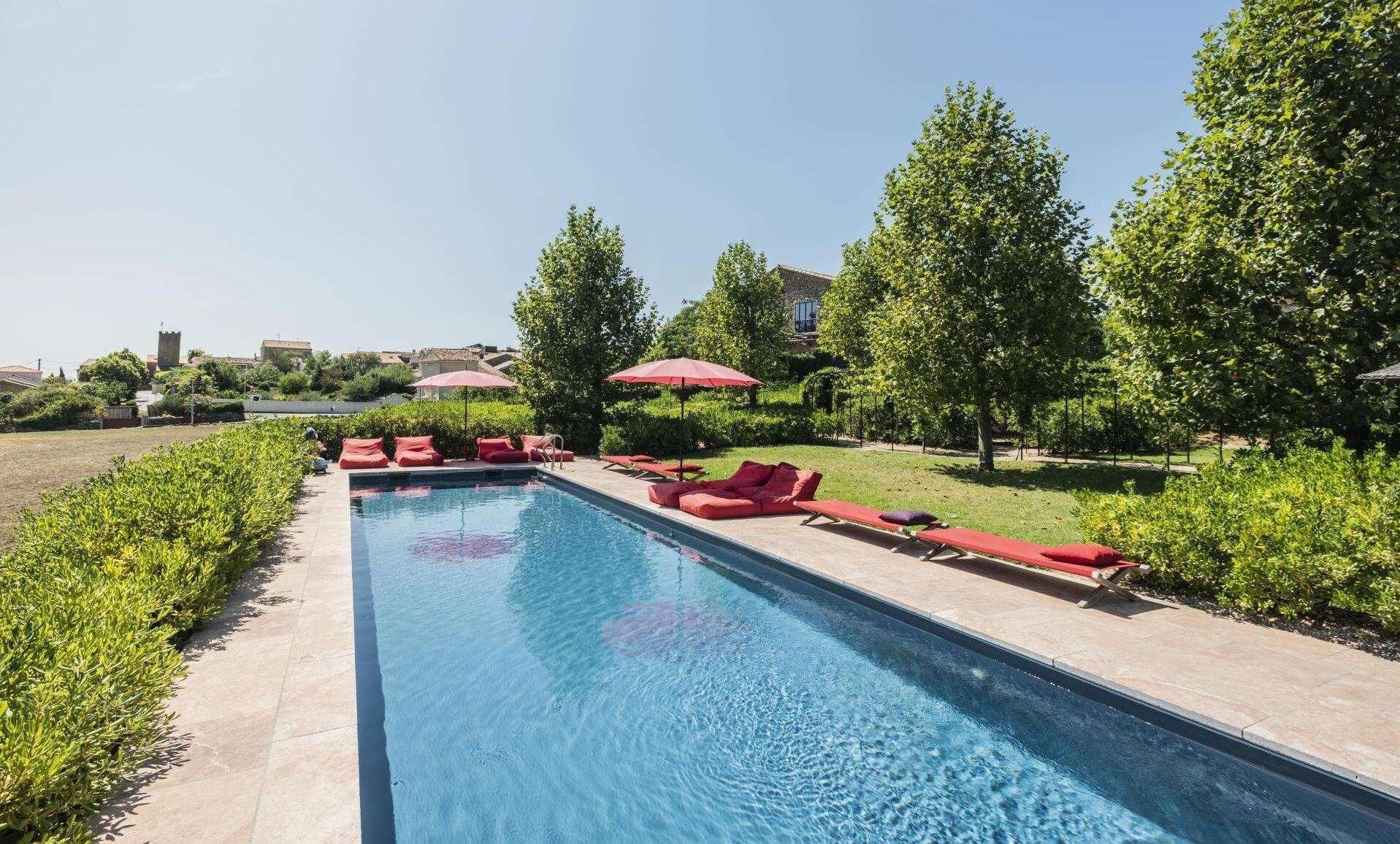 Una piscina con lettini e ombrelloni rossi, immersa nel verde e circondata da edifici.