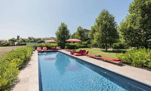 A swimming pool with red lounge chairs and umbrellas, surrounded by greenery and buildings. - Château & Village Castigno - Wine Hotel & Resort (Assignan)