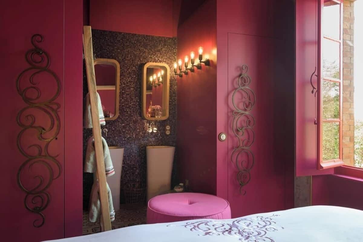 A room with a bed, a pink ottoman, a mirror with lights, and a window.