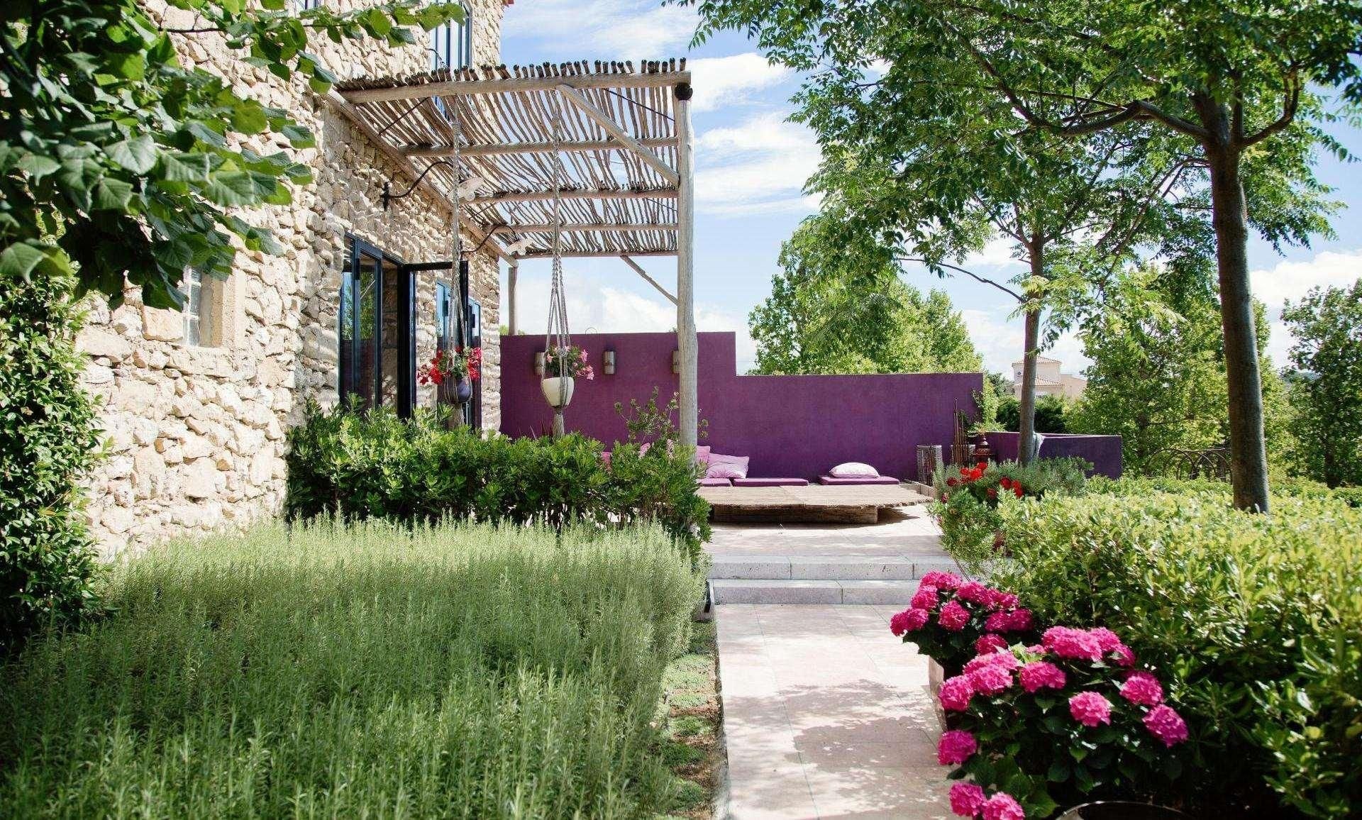 A stone building with a wooden pergola, a purple wall, and a garden with pink flowers.