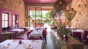 Un restaurante con paredes de piedra, detalles morados y lámparas colgantes. Las mesas están dispuestas con manteles blancos y sillas rosas.