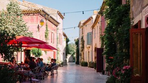 Una calle adoquinada con mesas al aire libre, edificios con contraventanas y luces colgantes.