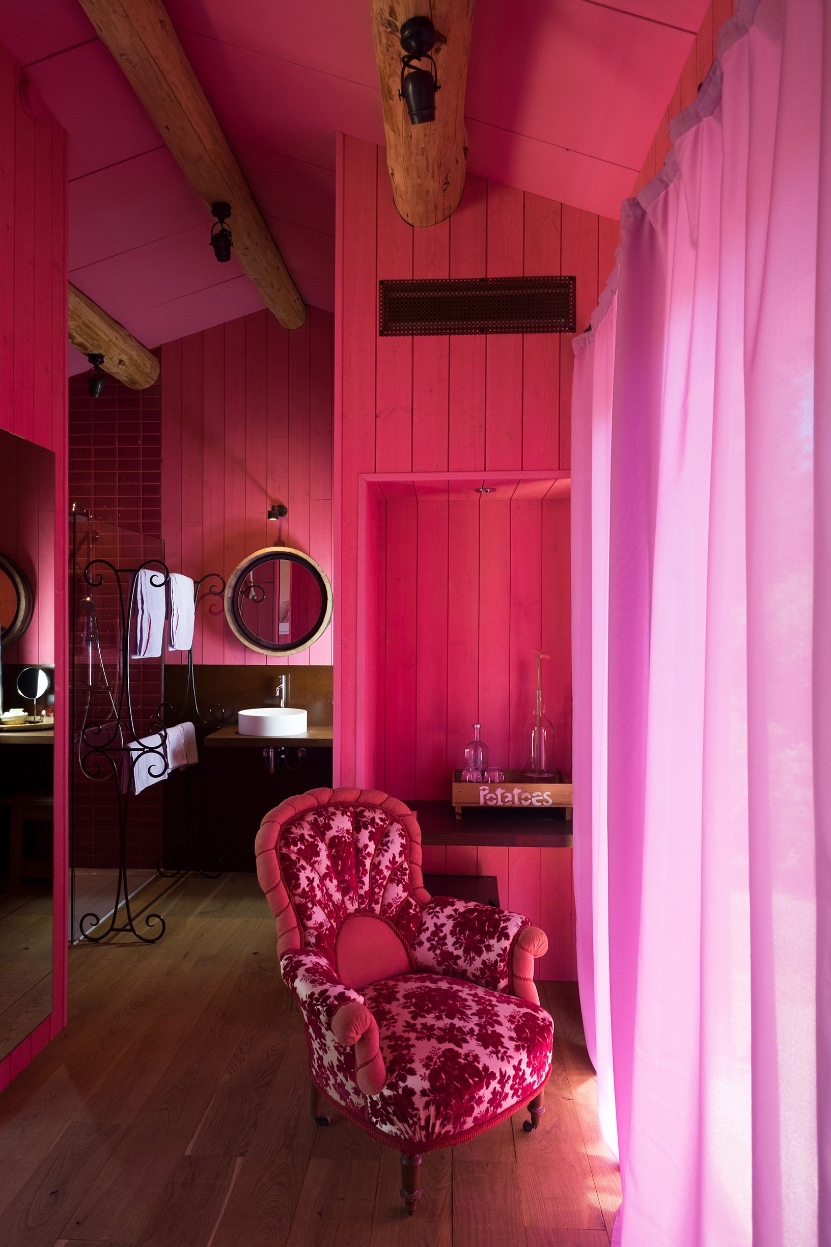 Una stanza in tonalità rosa con soffitto in legno, uno specchio tondo, un mobile toilette con lavabo e una poltrona con motivi floreali.