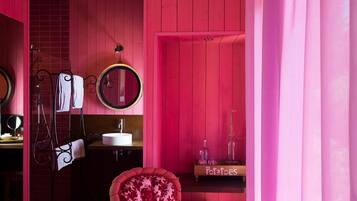 Una stanza in tonalità rosa con soffitto in legno, uno specchio tondo, un mobile toilette con lavabo e una poltrona con motivi floreali.