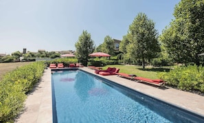 A swimming pool with red lounge chairs and umbrellas, surrounded by greenery and buildings.