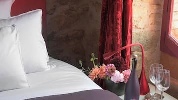 Uma cama com lençóis brancos, um vaso de flores, uma garrafa de vinho e duas taças em uma mesa de cabeceira.