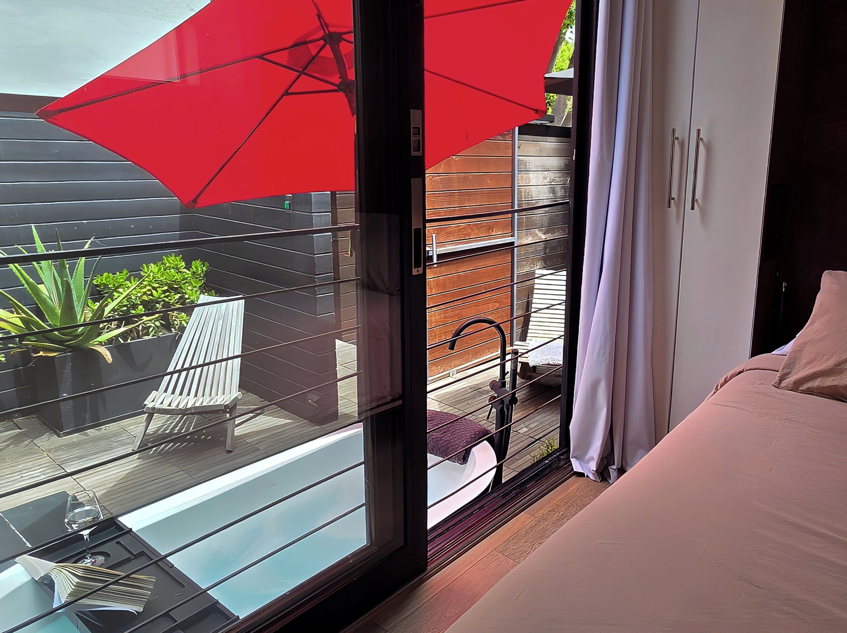 A modern bedroom with a large window, a red umbrella, and a view of a wooden deck with plants.