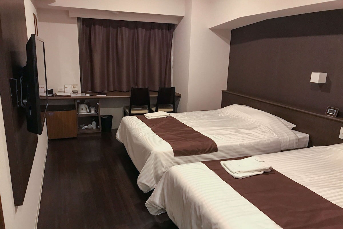 A hotel room with two beds, a desk with chairs, a television, and a window with curtains.