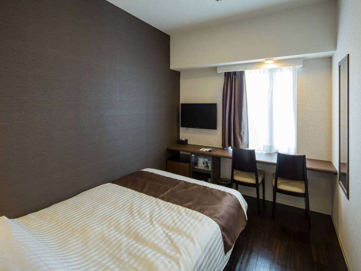 A hotel room with a bed, a desk with two chairs, a television, and a window with curtains.