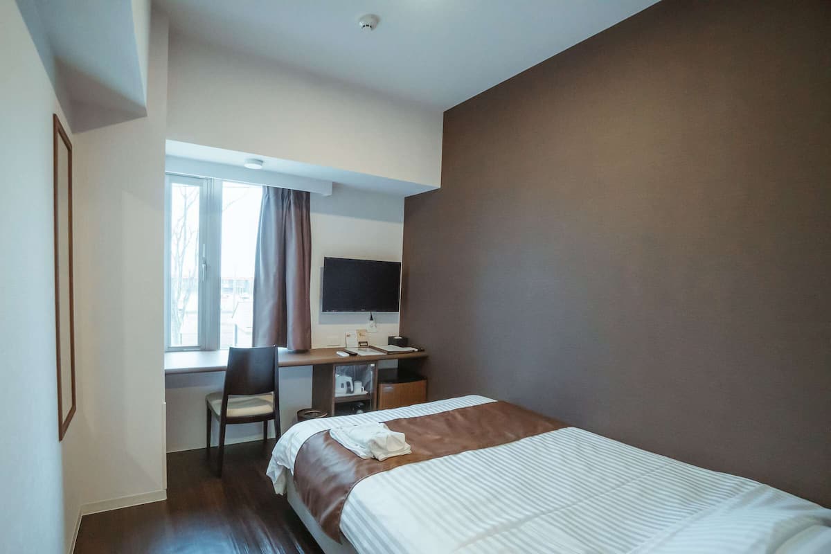 A hotel room with a bed, a desk with a computer and a chair, a window with curtains, and a flat-screen TV.