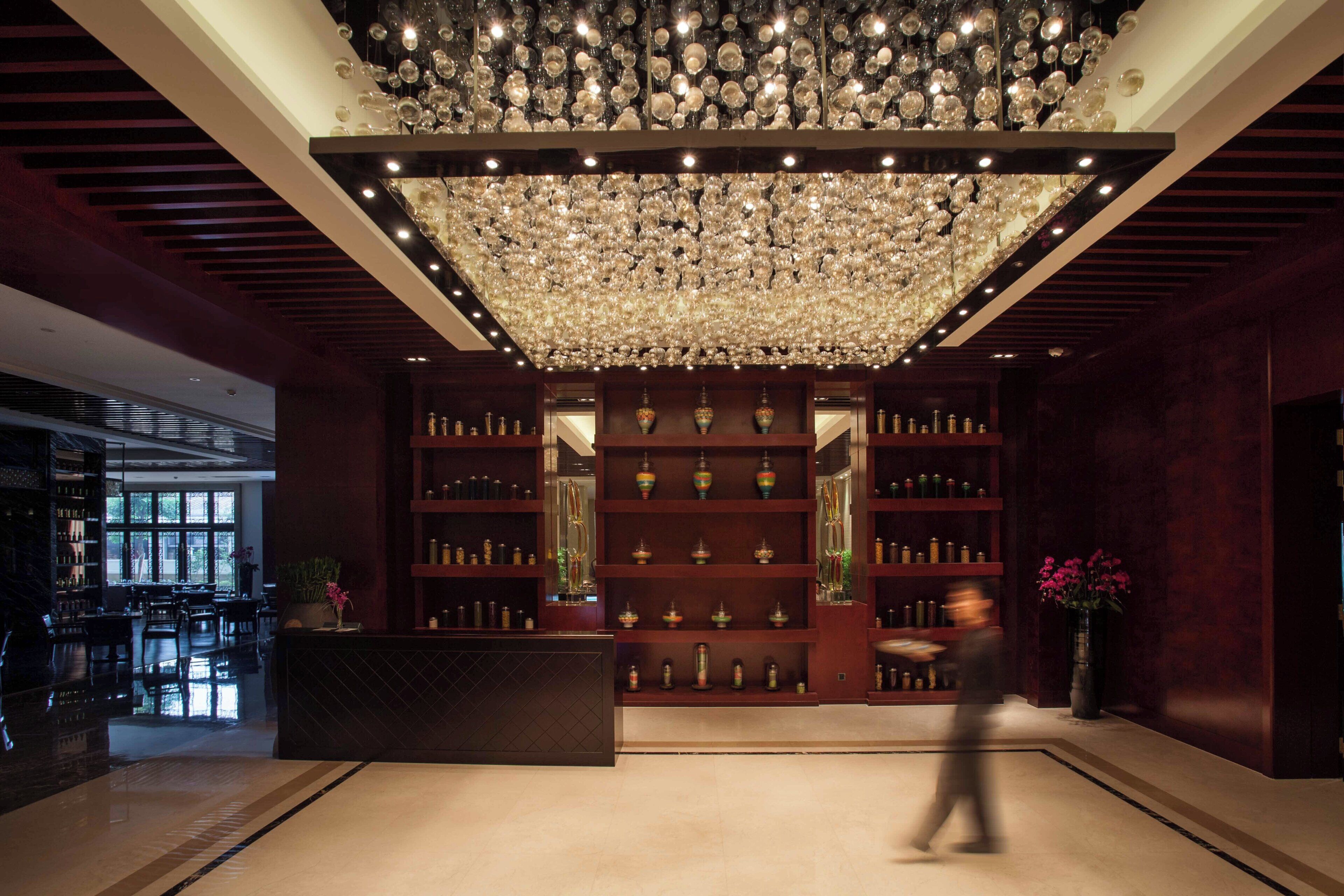 A lobby with a large chandelier, wooden shelving with decorative items, and a reception desk.