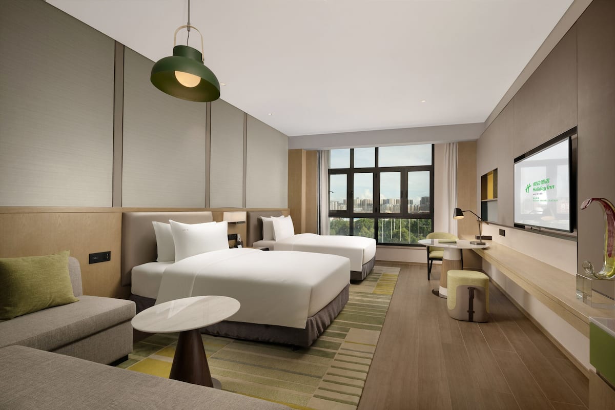 A modern hotel room with two beds, a sofa, a small table, and a large window offering a city view.