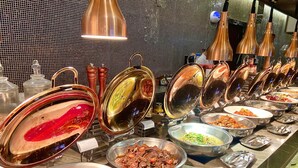 Un buffet proposant divers plats présentés dans des plats de service en métal, dont certains sont munis de couvercles, et un mur carrelé en arrière-plan.