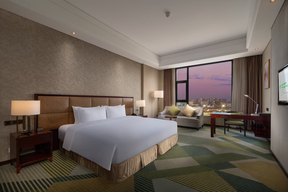 A hotel room with a large bed, a desk, a chair, and a view of the city at dusk.