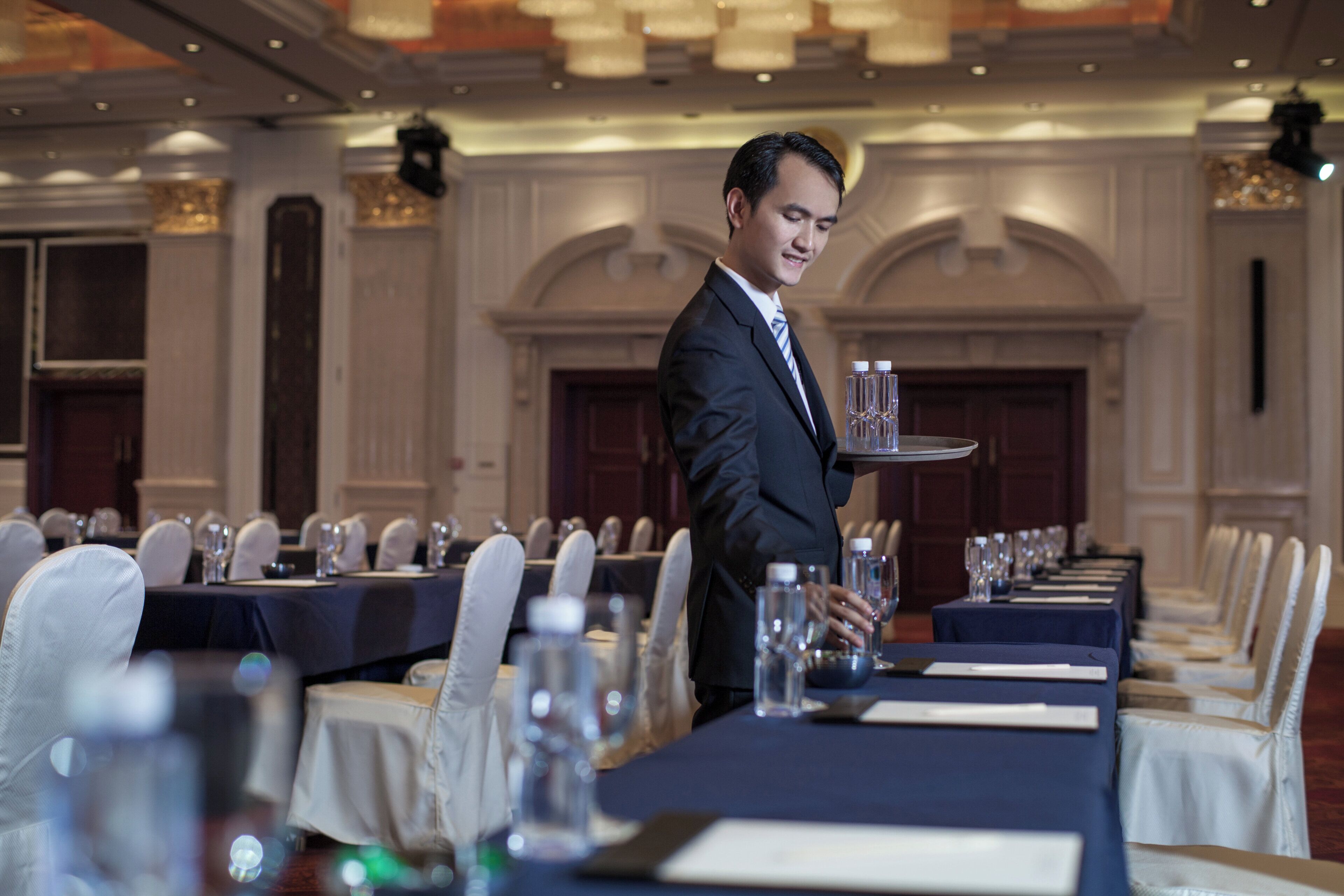 Un ambiente de comedor formal con un camarero llevando una bandeja con vasos y botellas de agua. Las mesas están dispuestas con manteles blancos, sillas y cristalería.