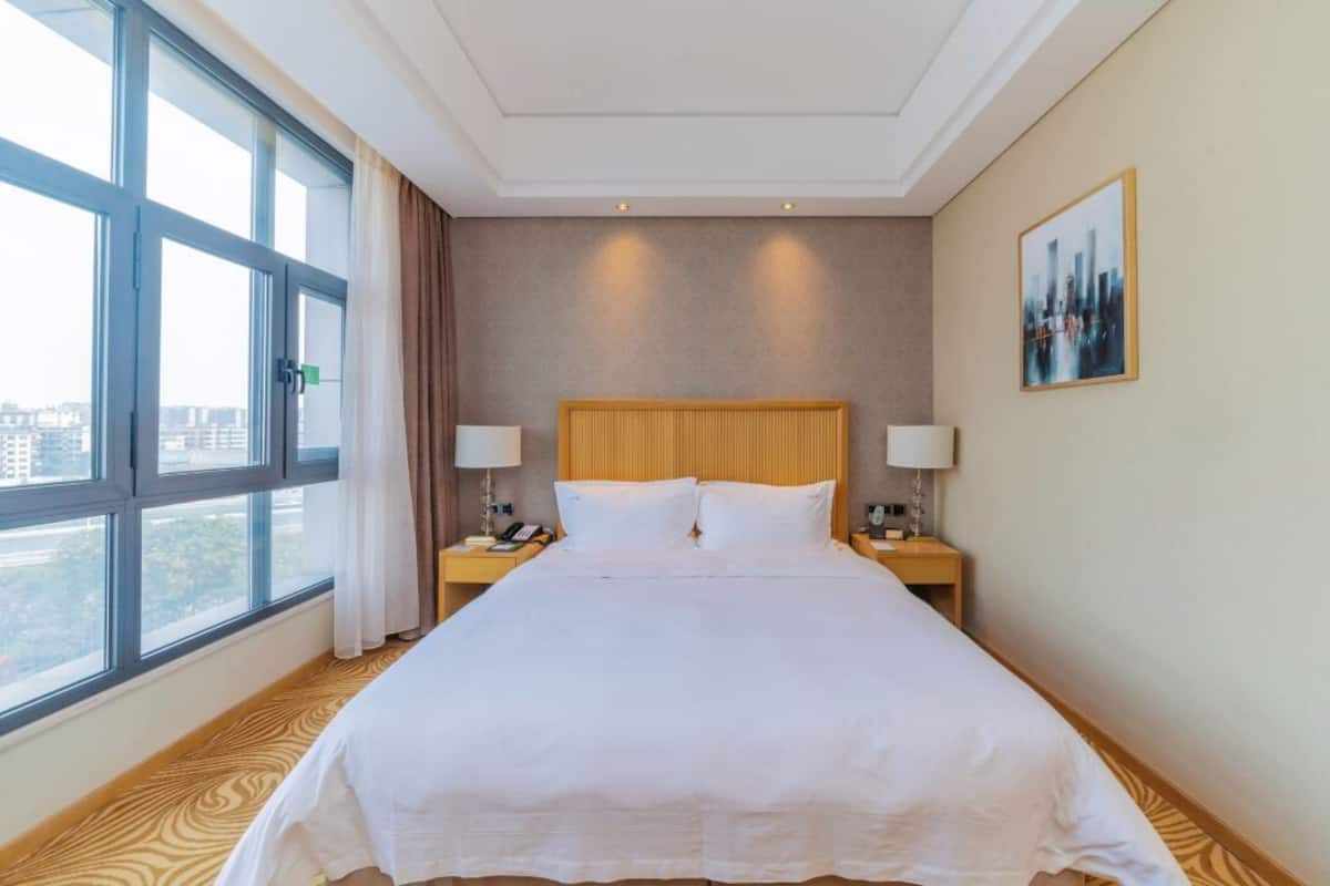 A hotel room with a large window, a bed with white bedding, two bedside tables with lamps, and a cityscape view.