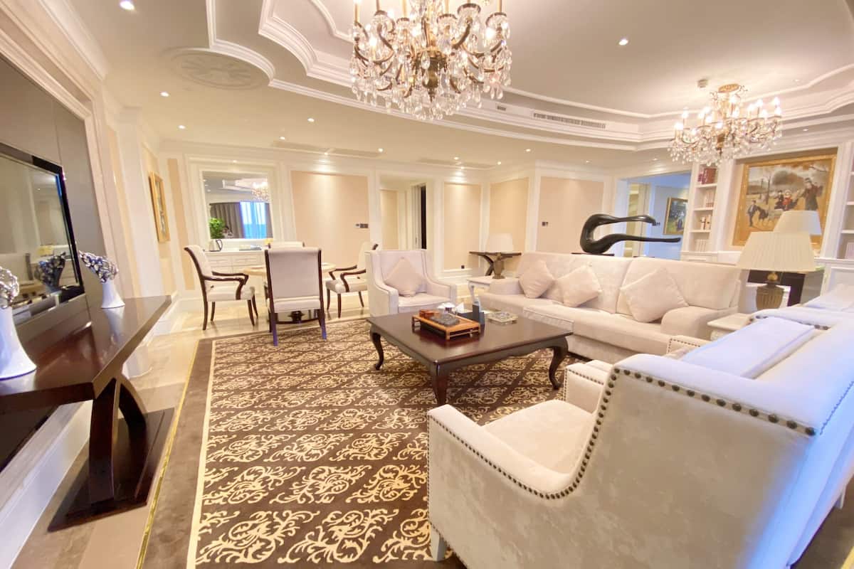 A spacious living room with a large white sofa, a coffee table, and a chandelier. There is a dining area with a table and chairs in the background.