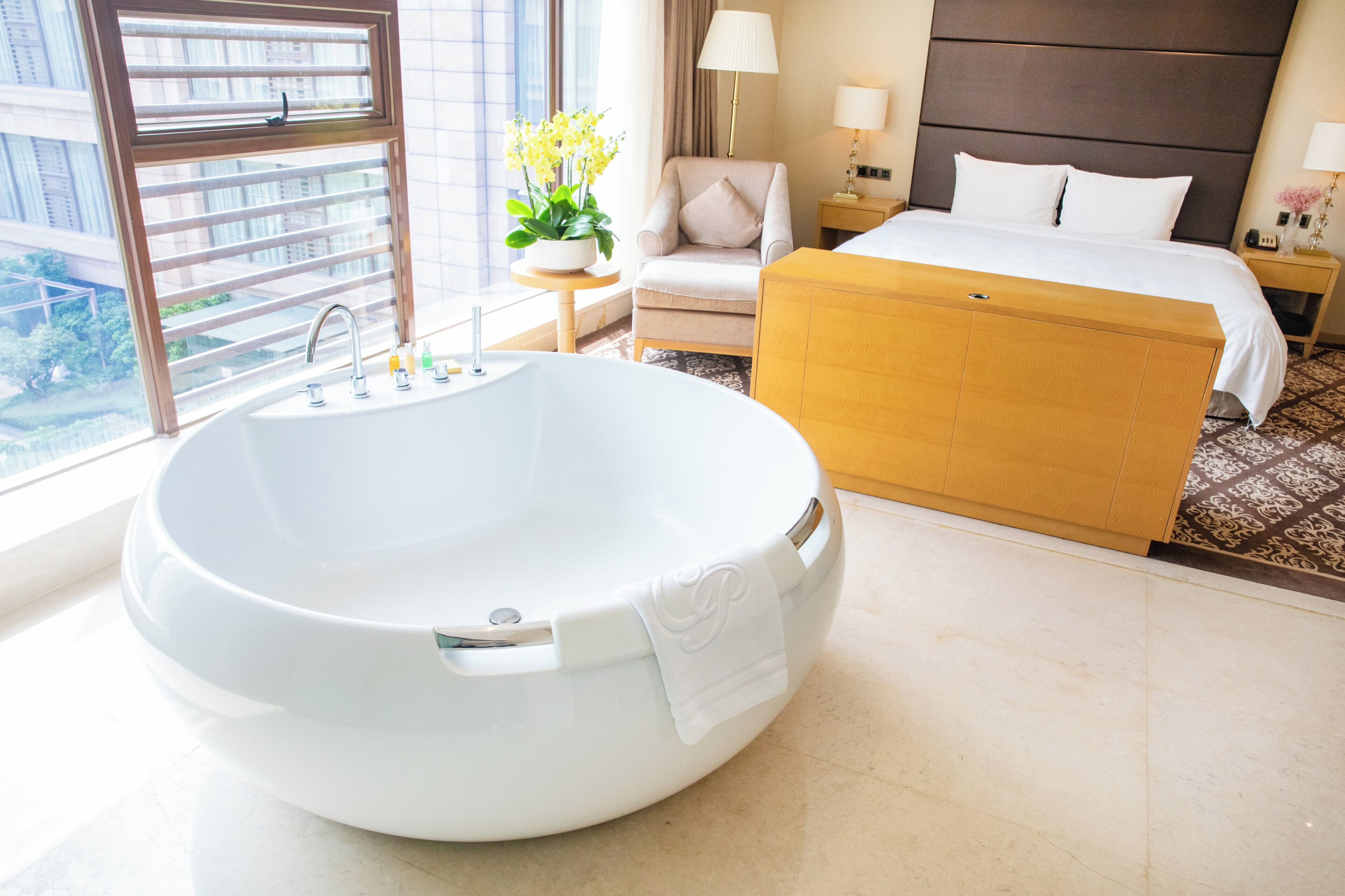 A modern hotel room with a large freestanding bathtub, a bed with white linens, a small round table with a lamp, and a view of the city through large windows.