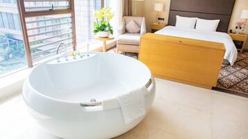 A modern hotel room with a large freestanding bathtub, a bed with white linens, a small round table with a lamp, and a view of the city through large windows.