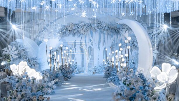 Un espace intérieur doté d’une allée centrale menant à une arche décorée, entourée de compositions florales et illuminée par des guirlandes lumineuses suspendues.
