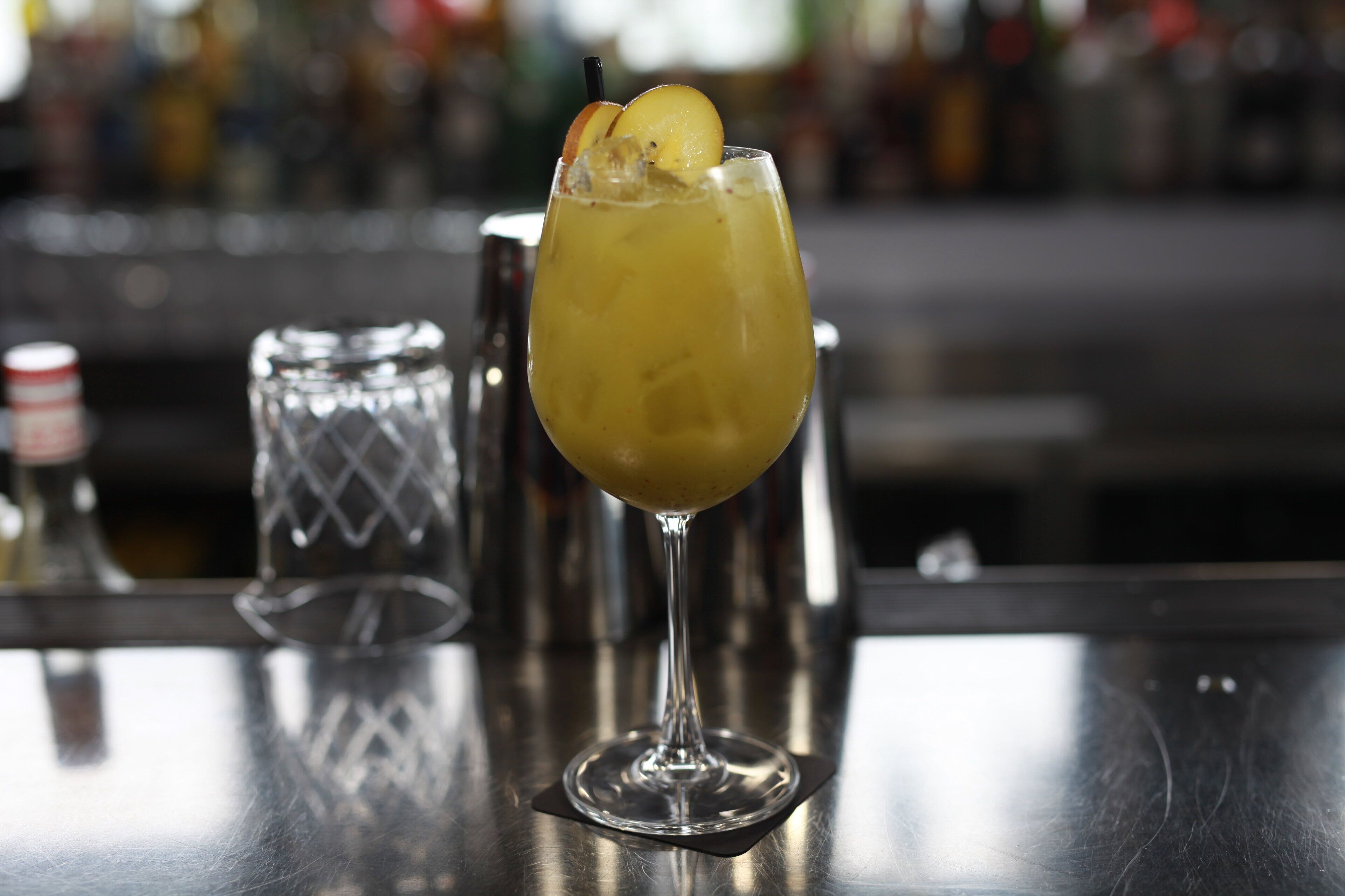 A cocktail in a glass with an orange slice garnish, placed on a bar counter with a blurred background of bottles.