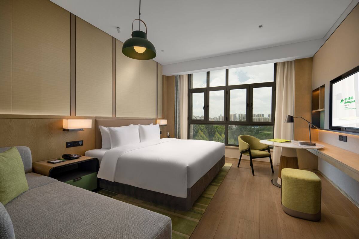 A modern hotel room with a large bed, a desk, a chair, and a view of the city through large windows.