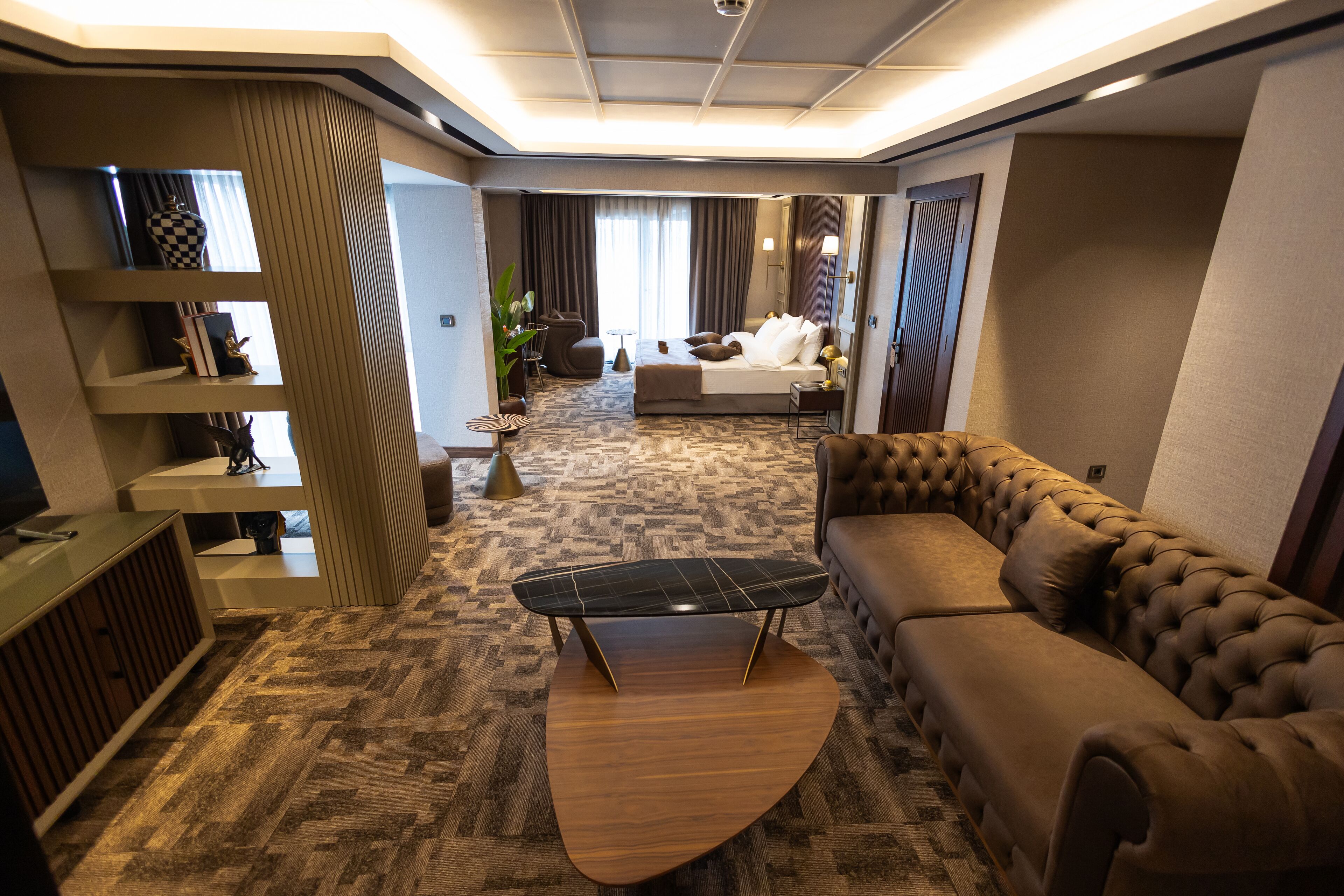 A modern hotel room with a large bed, a leather sofa, a unique coffee table, and a built-in TV.
