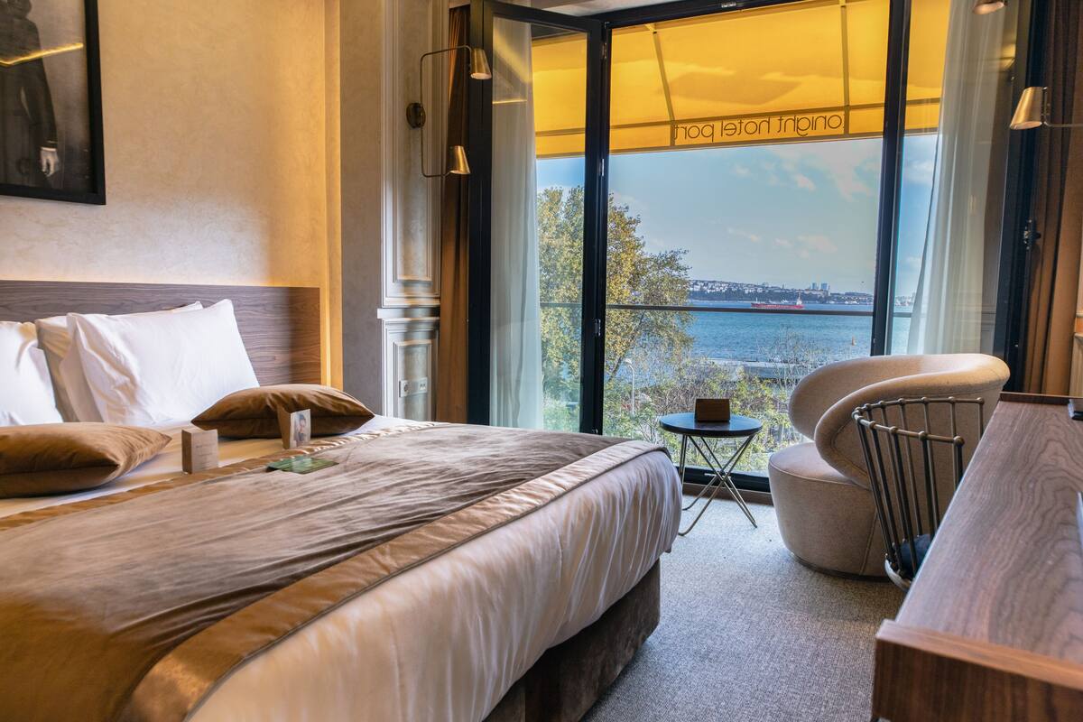 A hotel room with a large bed, a small round table, a chair, and a view of a body of water through large windows.