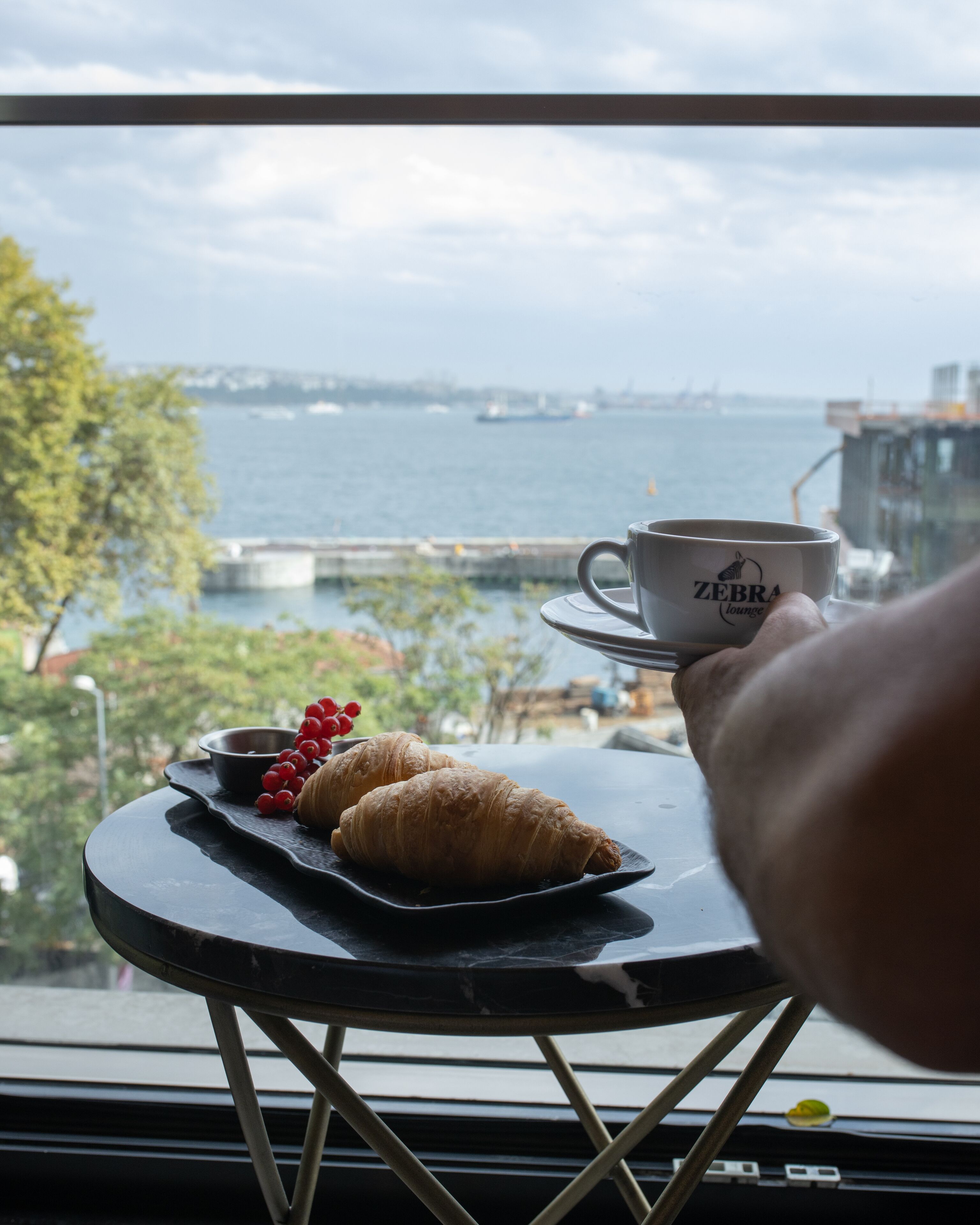 Ein Tisch mit einer Tasse und Gebäck, Blick aus dem Fenster auf ein Gewässer und entfernte Gebäude.