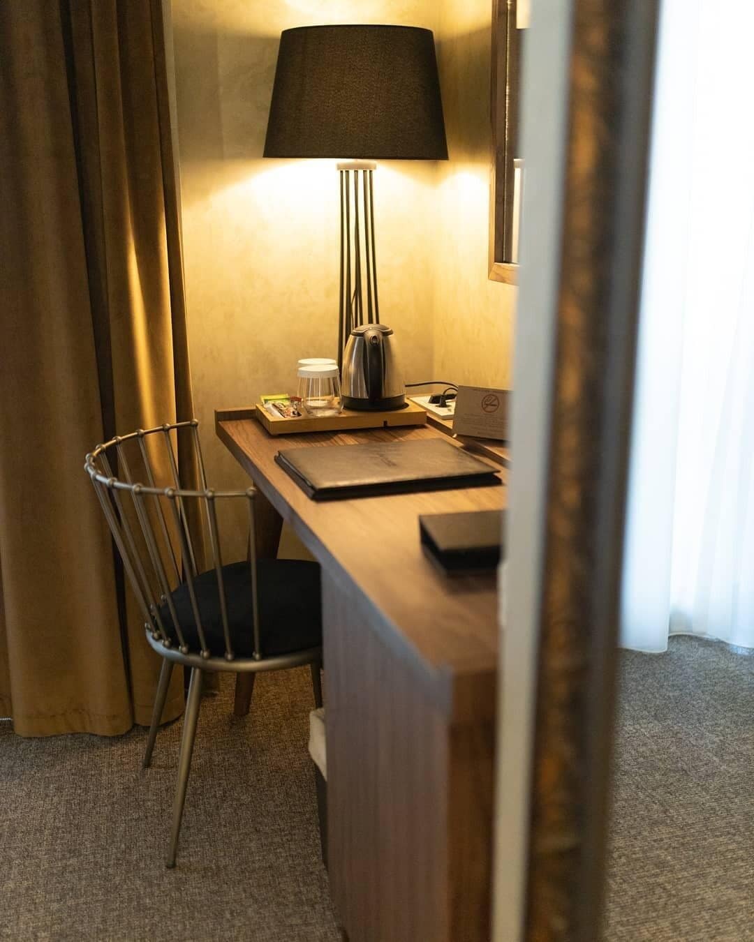 A hotel room with a wooden desk, a metal chair, a table lamp, a kettle, a cup, and a notebook.