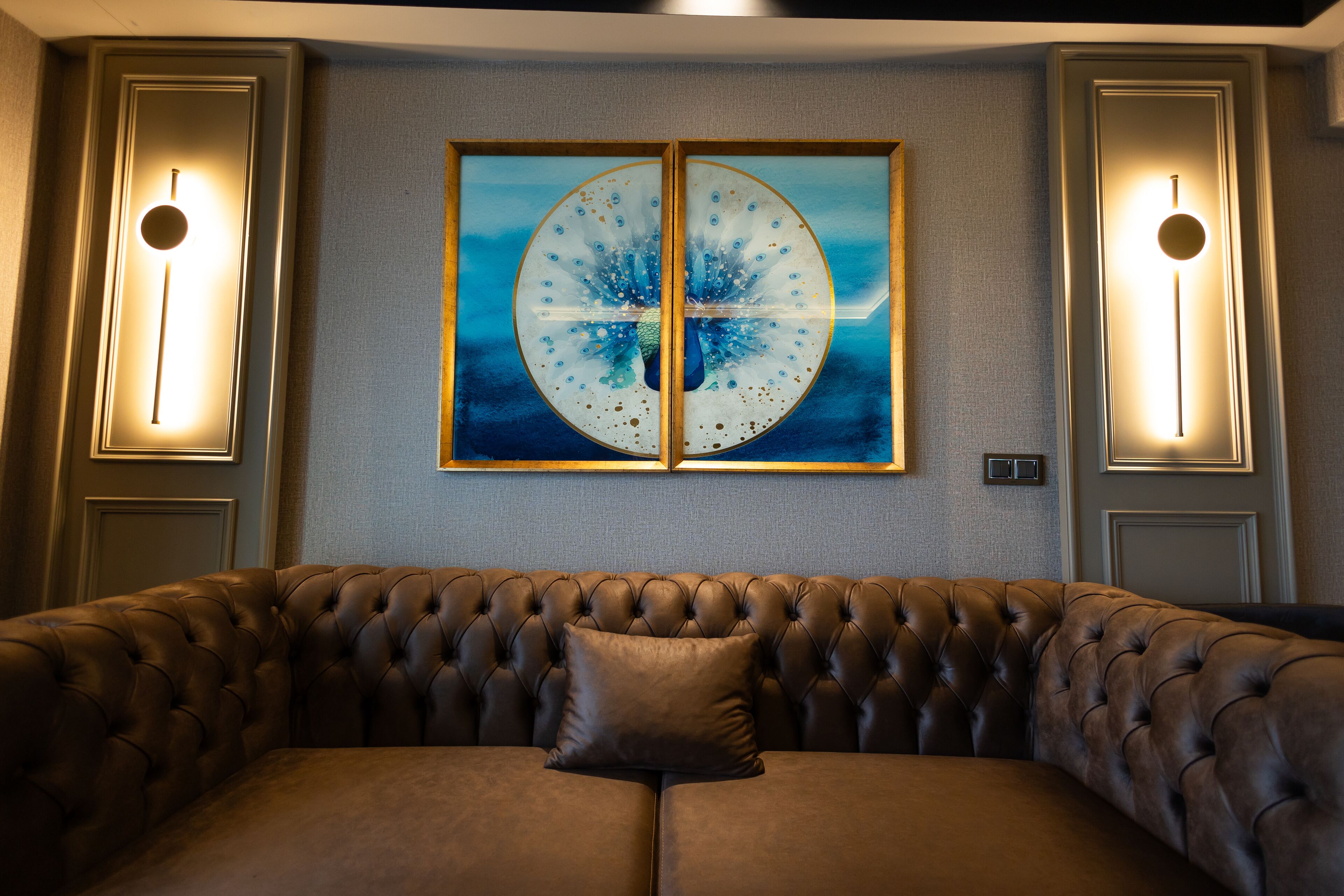 A tufted leather sofa with a single pillow, a framed artwork with a circular design, and wall sconces on either side.