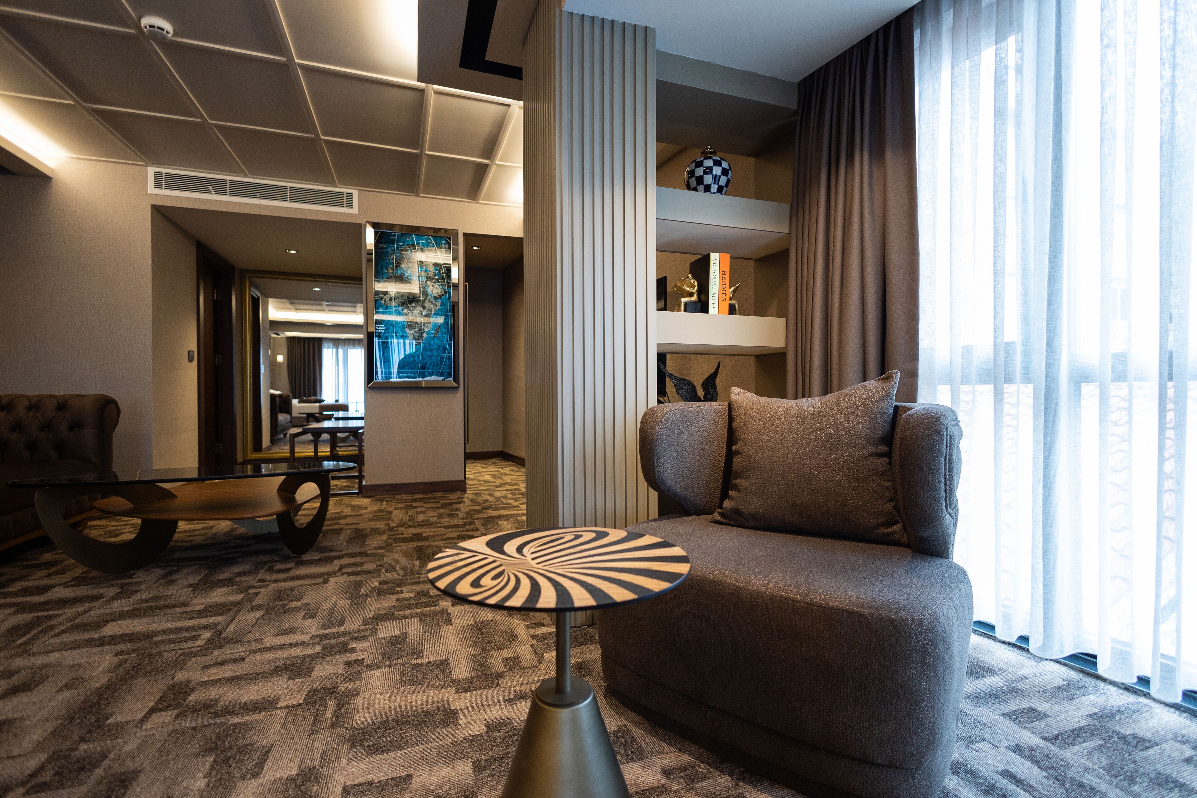 A modern hotel room with a sofa, a unique table lamp, a large window with curtains, and a view of the city.