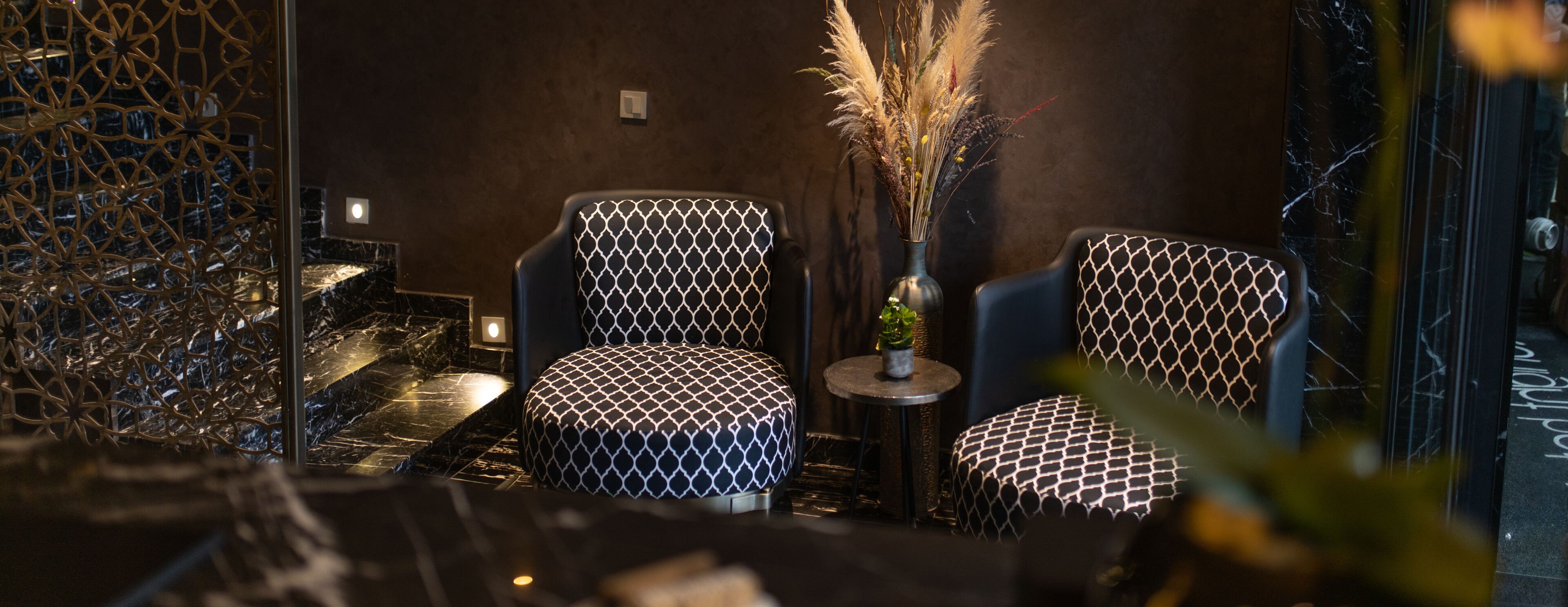 Ein modernes Interieur mit zwei gemusterten Stühlen, einem kleinen Tisch mit einer Vase getrockneter Pflanzen und einer dekorativen Wand.