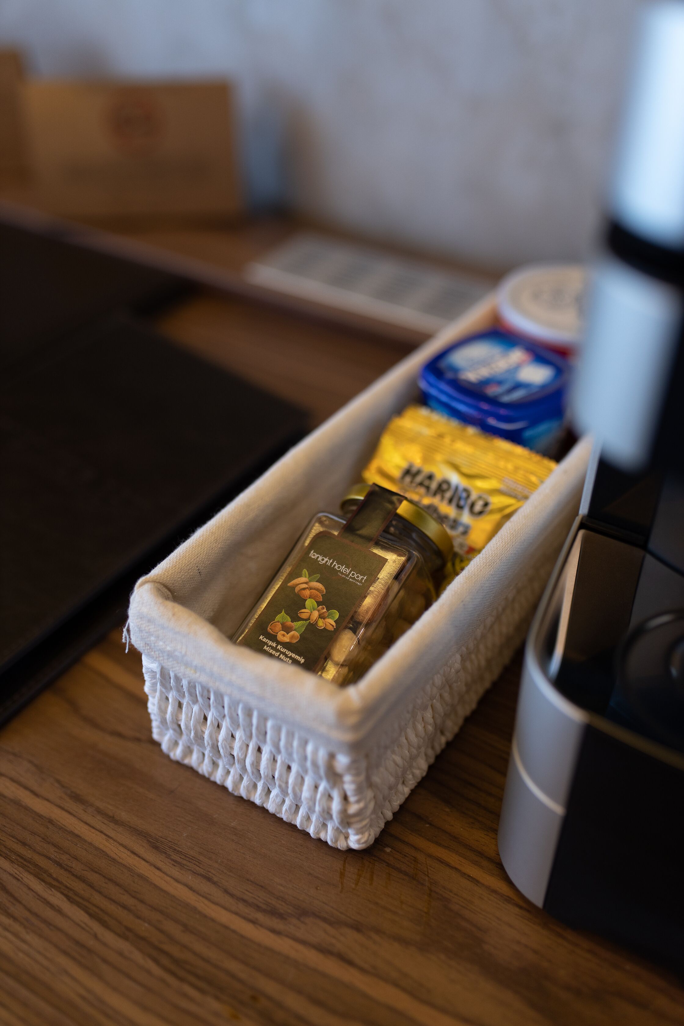 A small woven basket containing a bottle of Haribo fruit gum, a jar of honey, and a blue container on a wooden table.
