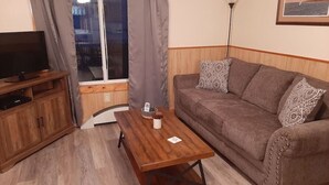 A living room with a wooden coffee table, a brown sofa with patterned cushions, a TV on a wooden stand, and a window with curtains.