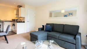 A modern living room with a grey sectional sofa, a glass coffee table, and a kitchen area in the background. - No16 (Mansfield)