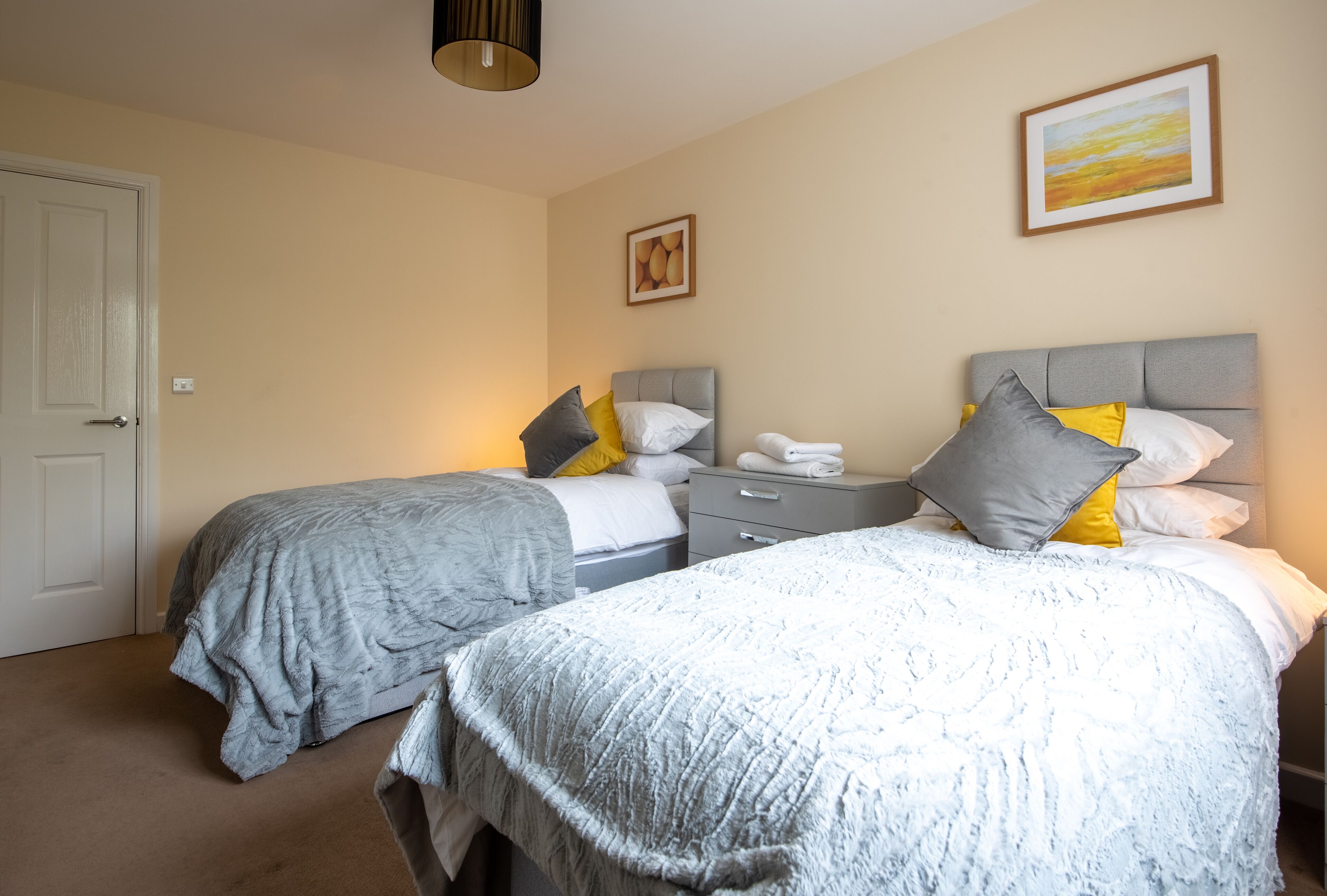 A bedroom with two beds, a bedside table, a nightstand, a lamp, and a painting on the wall.