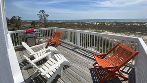 Um terraço com duas cadeiras Adirondack brancas e duas cadeiras Adirondack laranja, com vista para uma praia com dunas e o oceano.