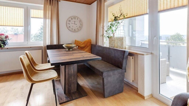 Een moderne woonkamer met een houten plafond, een eettafel met stoelen, een bank en een groot raam met jaloezieën.