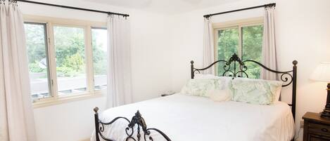 A bedroom with a bed, a ceiling fan, two windows with curtains, and a nightstand with a lamp.