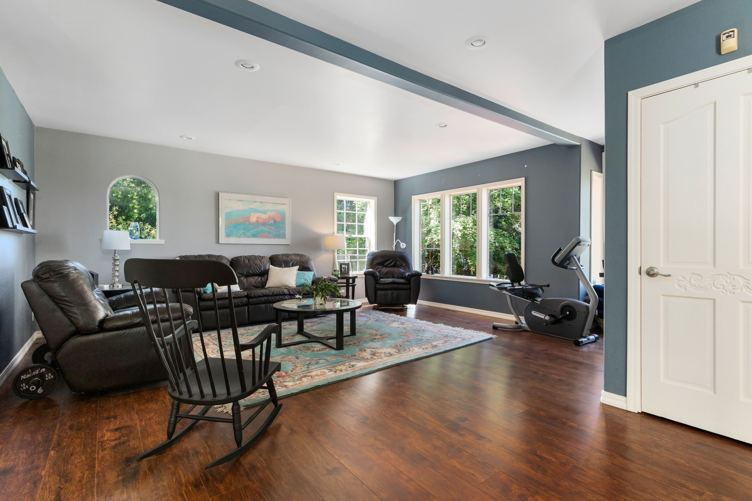 A spacious living room with a dark wood floor, a black leather sofa, a rocking chair, and a large window providing natural light.