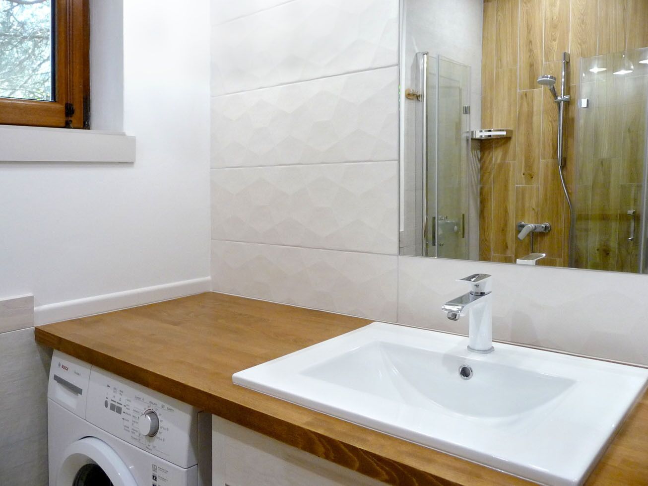A bathroom with a wooden countertop, a white sink, a washing machine, and a glass shower enclosure.