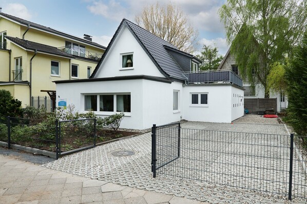 Una casa moderna de dos plantas con fachada blanca, techo gris y un jardín delantero cercado.