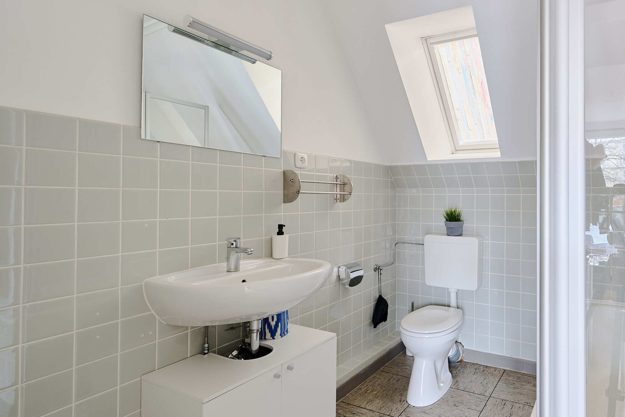 Une salle de bain moderne avec un lavabo blanc, un miroir et des toilettes. Une lucarne se trouve au-dessus des toilettes et une petite plante est posée sur le réservoir.
