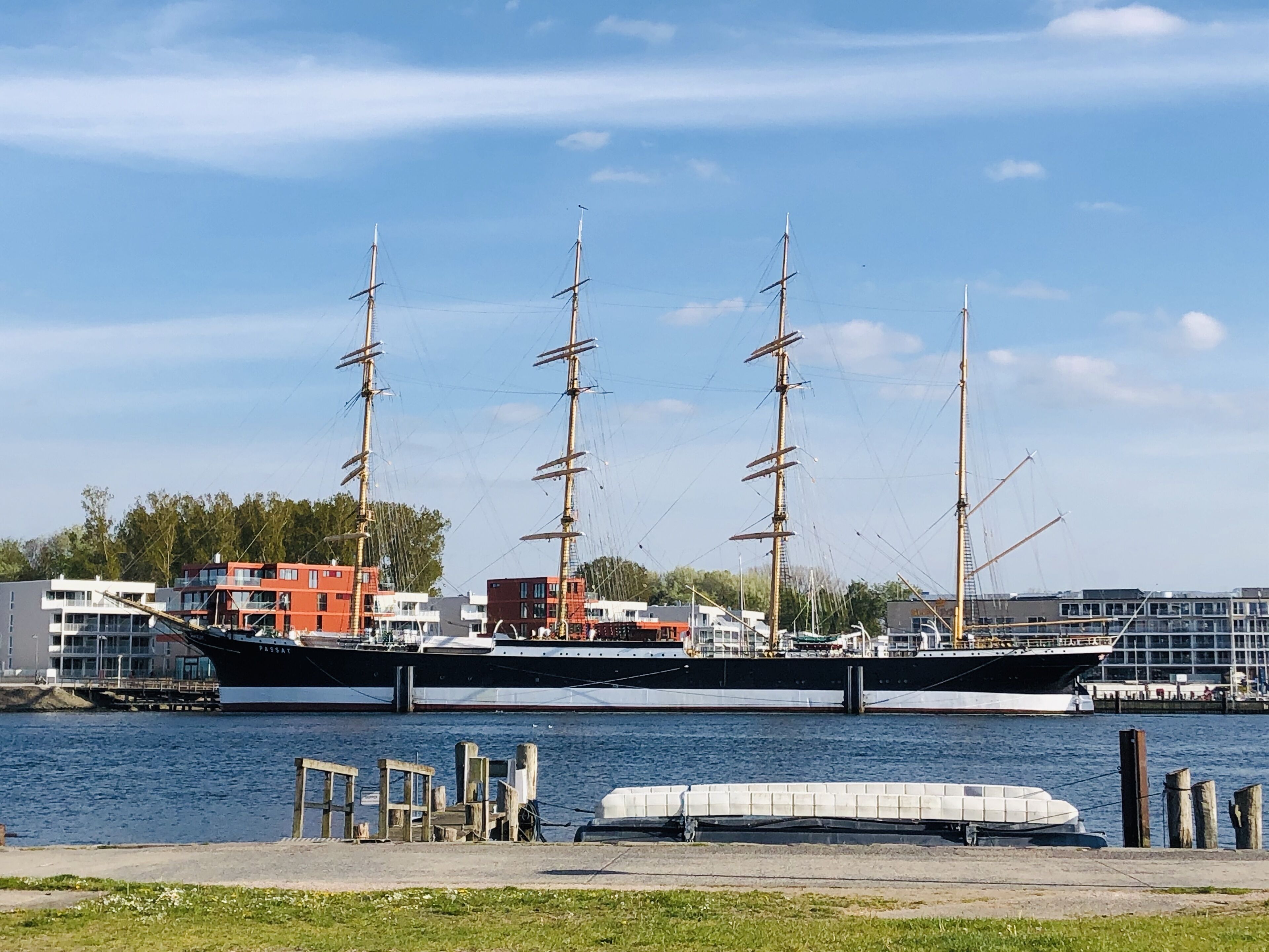 Ett stort segelfartyg låg förtöjt vid en småbåtshamn, med moderna byggnader i bakgrunden.