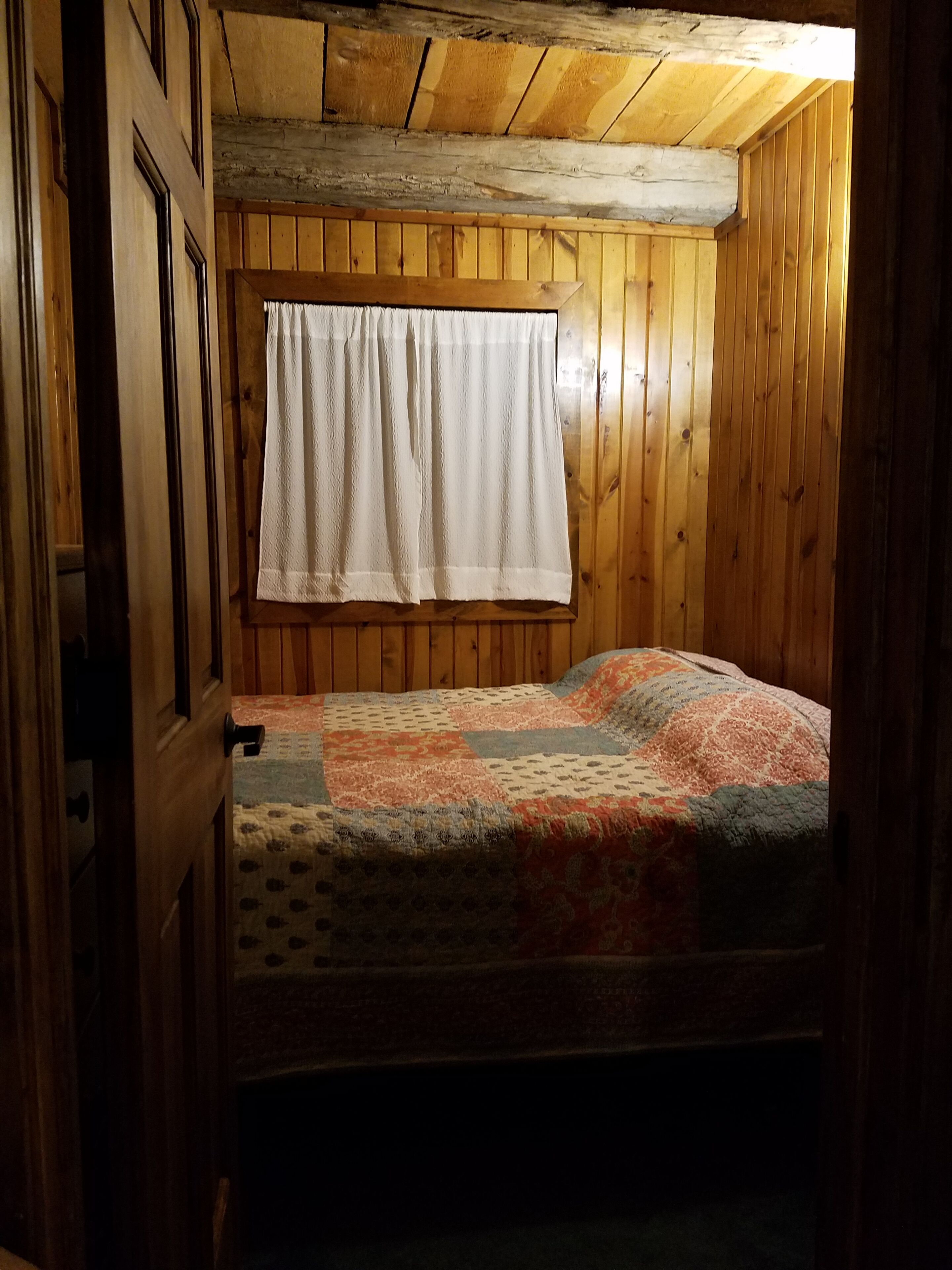 Une petite pièce en bois, aménagée comme une cabane, avec un lit simple, une fenêtre ornée d’un rideau blanc et une porte en bois.