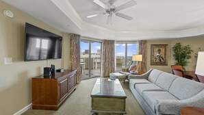 A modern living room with a flat-screen TV, a wooden cabinet, a glass coffee table, a grey sofa, and a ceiling fan.