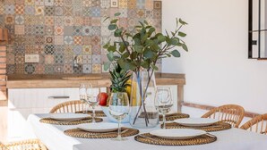 A dining table set with white plates, woven placemats, and wine glasses, accompanied by a vase with green foliage and a pineapple.