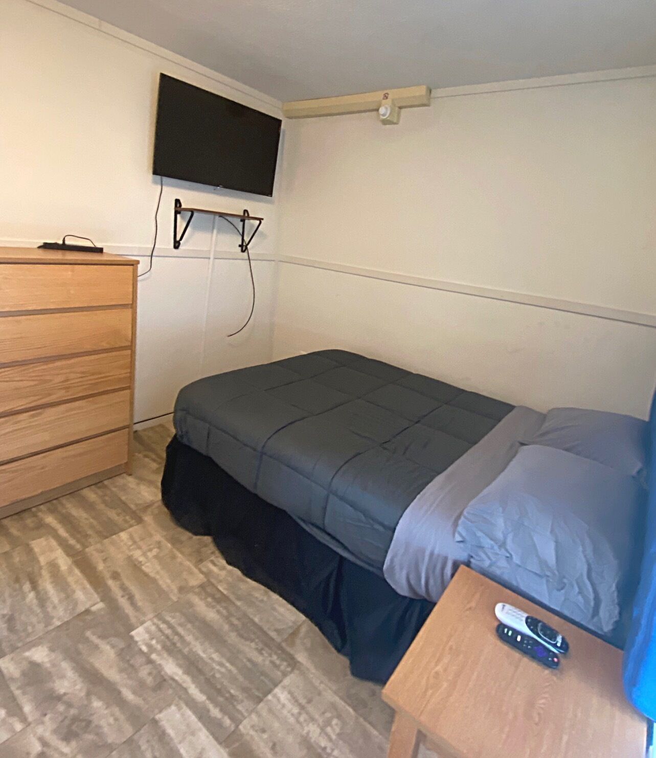 Une petite pièce, simplement aménagée, avec un lit, une commode en bois et un téléviseur fixé au mur.